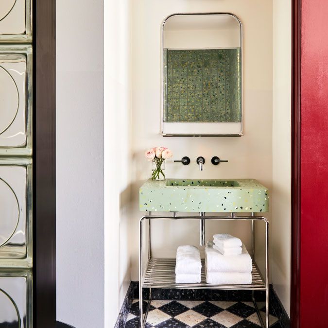 A stylish bathroom with a green terrazzo sink, a rectangular mirror, black and white checkered floor, and red wall. Towels are neatly stacked below.