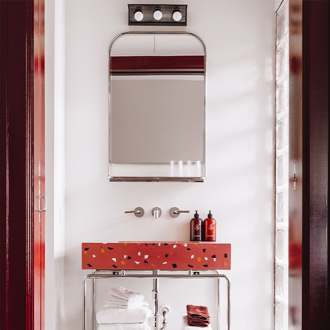 Bathroom with a red terrazzo sink, large mirror, wall-mounted faucet, and neatly stacked towels on a metal shelf below.
