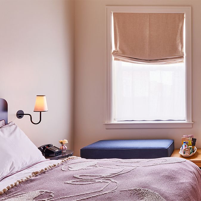 Cozy bedroom with a made bed, wall-mounted lamp, window with beige Roman shade, and a bedside table with a phone and decor items.