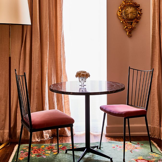 Cozy room with a round table, two pink chairs, ornate clock, and floral rug, framed by pink curtains and soft light from a window.