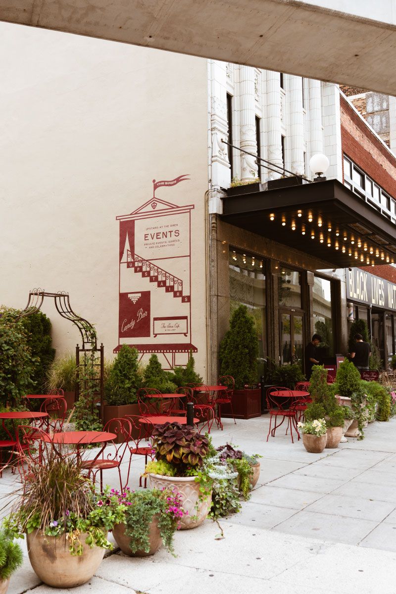 Outdoor cafe with red chairs and tables, surrounded by potted plants. A wall mural advertises events and candy bar inside.