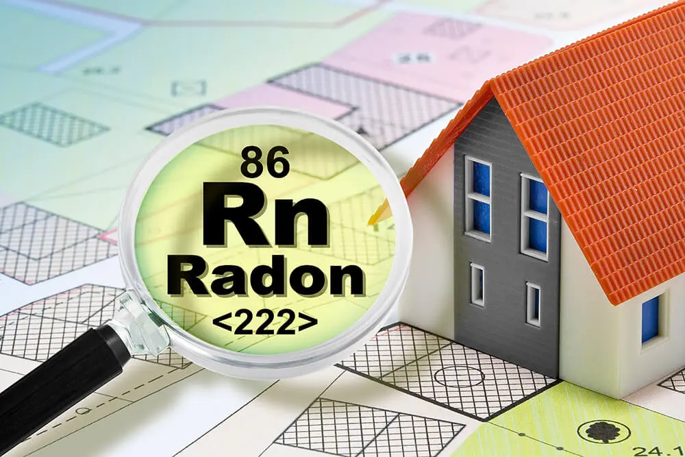 Modell eines Hauses auf einem Bauplan mit einer Lupe, die das chemische Symbol für Radon vergrößert, als Hinweis auf Radonsanierung