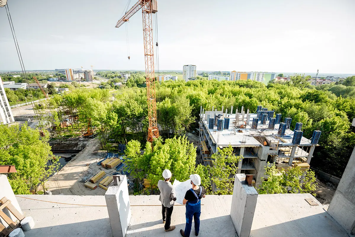 Bauplaner und Bauarbeiter stehen auf einem Rohbau und betrachten ein nachhaltiges Bauprojekt mit grünen Bäumen und Gebäuden im Hintergrund