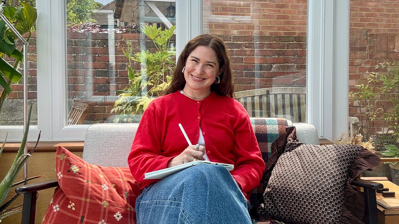 A person in a red sweater, sitting on a cushioned chair in a sunlit room, writing on a tablet with a stylus.