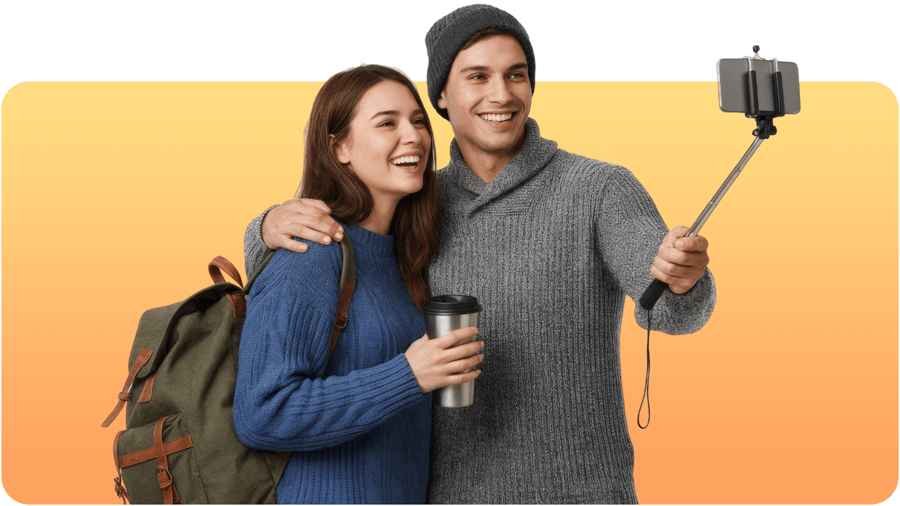 A smiling couple takes a selfie with a smartphone on a stick. The woman holds a backpack and a tumbler, and the man wears a beanie and sweater.