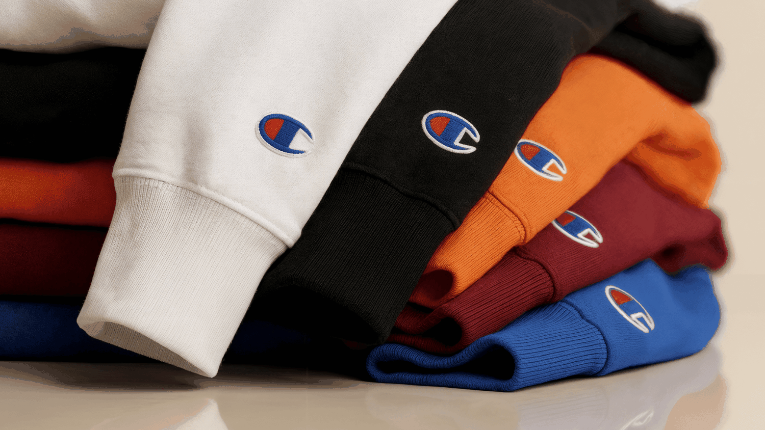 Folded colorful sweatshirts (white, black, orange, maroon, blue) stacked, showing embroidered Champion "C" logos on cuffs.