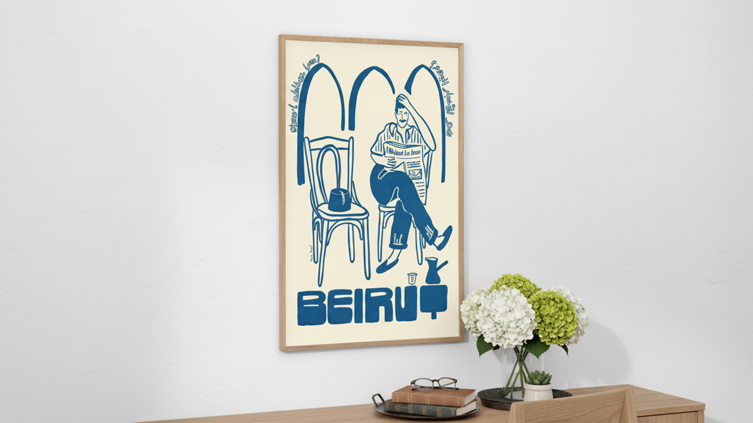 Framed poster of a person reading a newspaper in a café setting with the word "BEIRUT" below, on a wall above a wooden shelf with flowers and books.