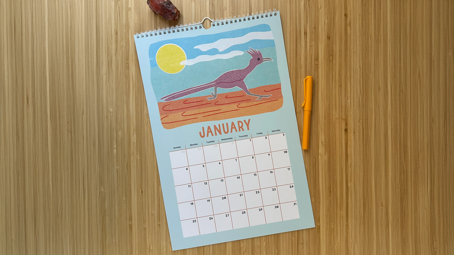 A wall calendar open to January, featuring an illustration of a bird on a desert landscape. A yellow pen is placed beside it on a wooden surface.