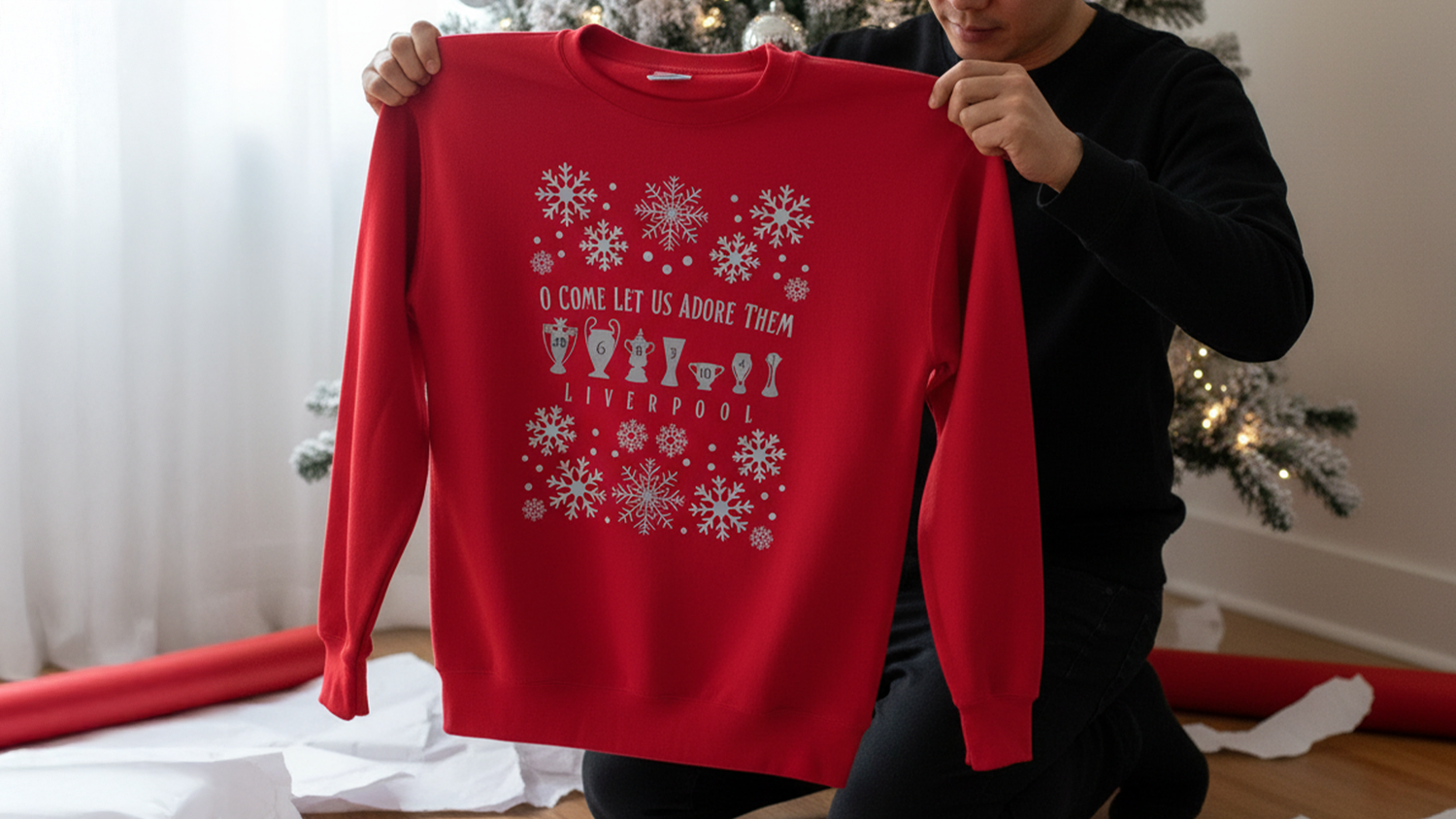 Person holding a red holiday sweater with festive drink icons and snowflakes, reading "O Come Let Us Adore Them, Liverpool," near a Christmas tree.