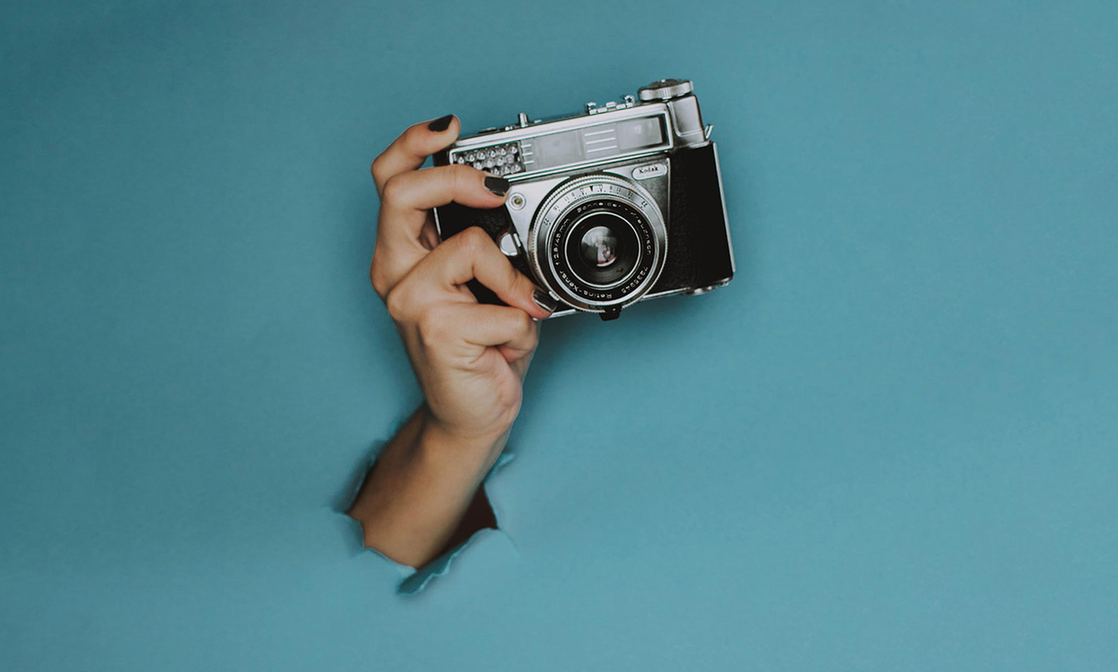 Eine Hand, die eine Vintage-Kamera hält, ragt durch einen zerrissenen blauen Papierhintergrund.