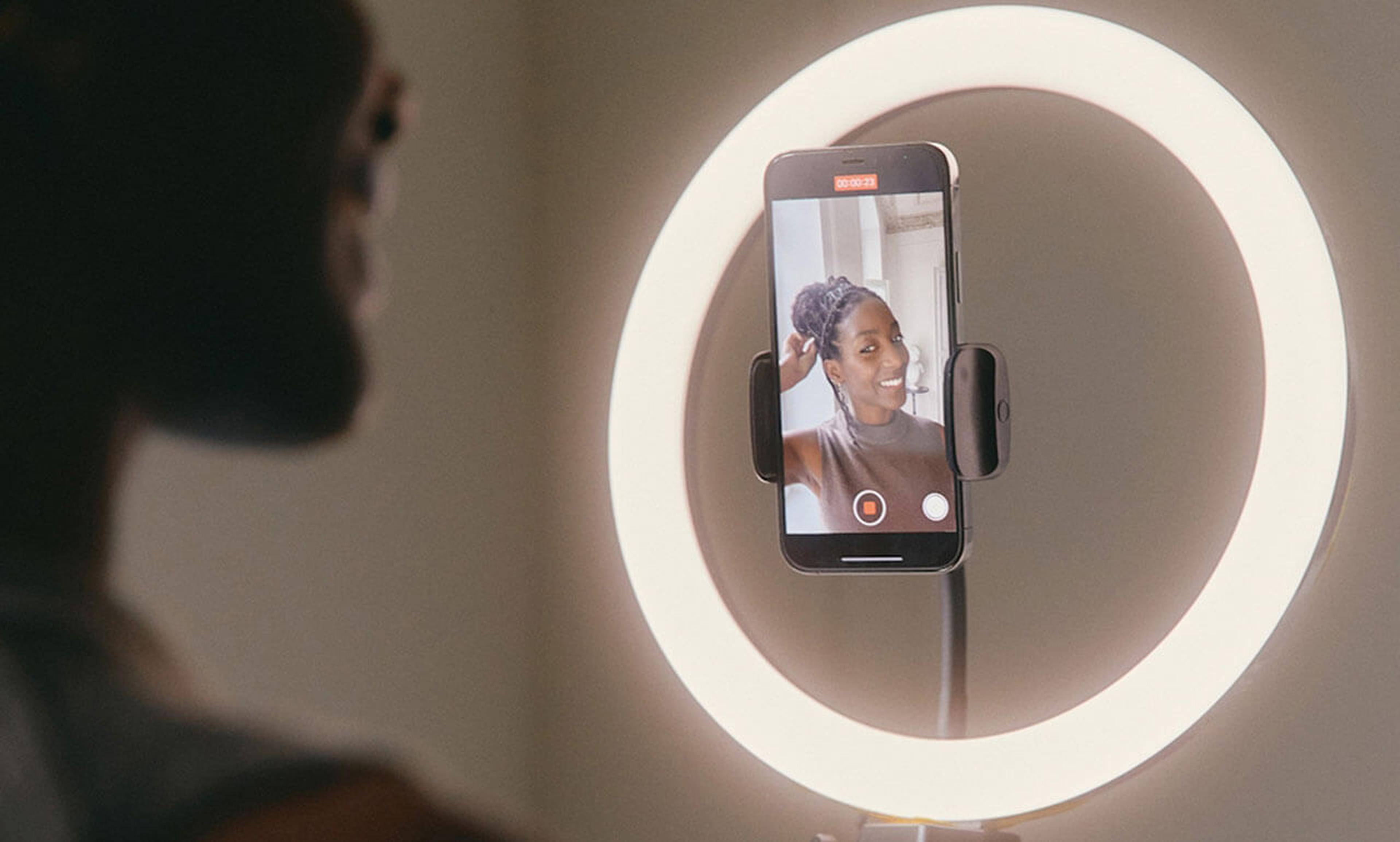 Person, die ein Video mit einem Smartphone aufnimmt, das in einem Ringlicht befestigt ist. Auf dem Bildschirm des Telefons ist ihr Bild zu sehen, was auf ein Selfie oder einen Video-Chat hindeutet.