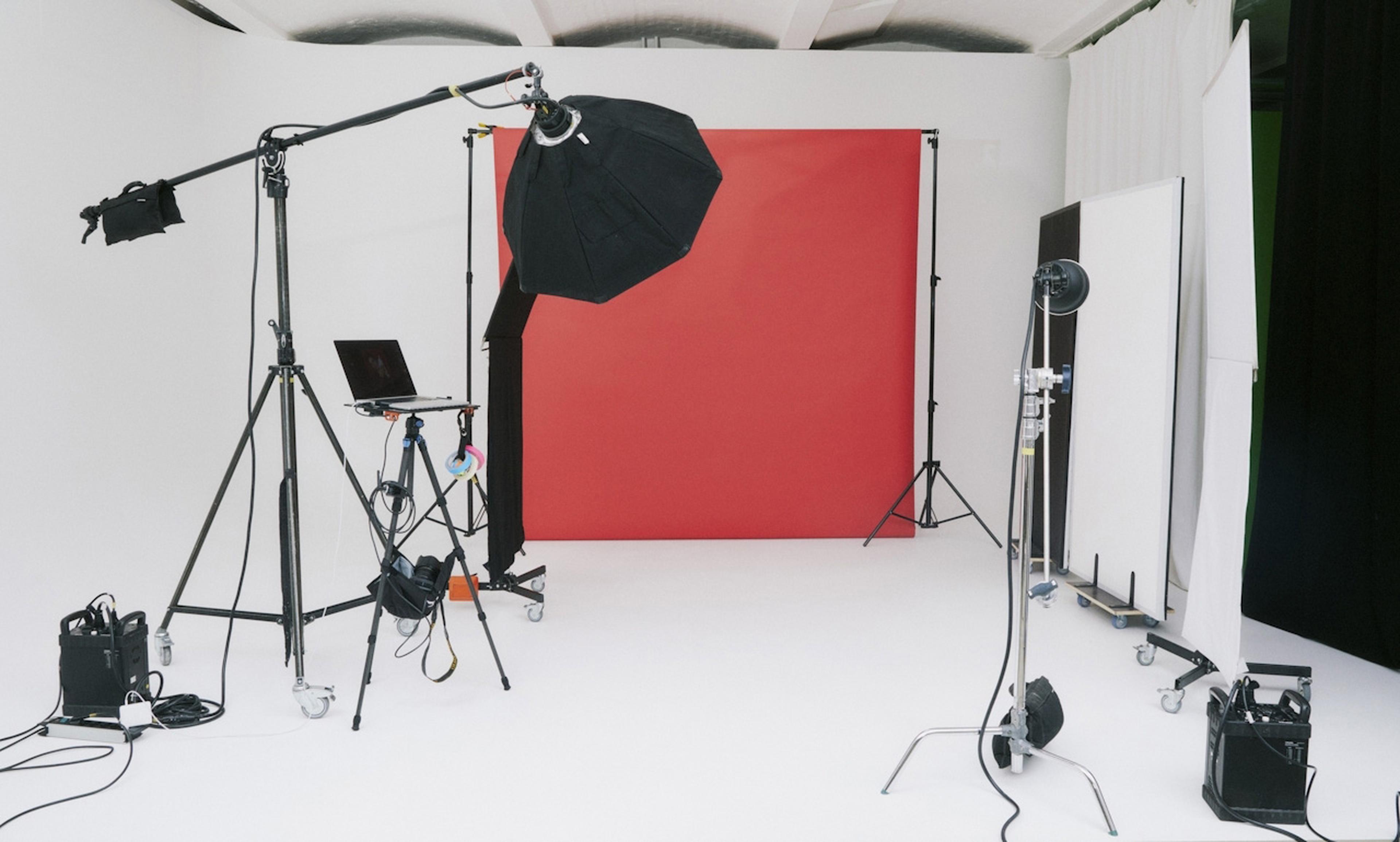 Fotostudio mit rotem Hintergrund, Beleuchtungsausrüstung, Stativ und Laptop-Aufbau auf weißem Boden.