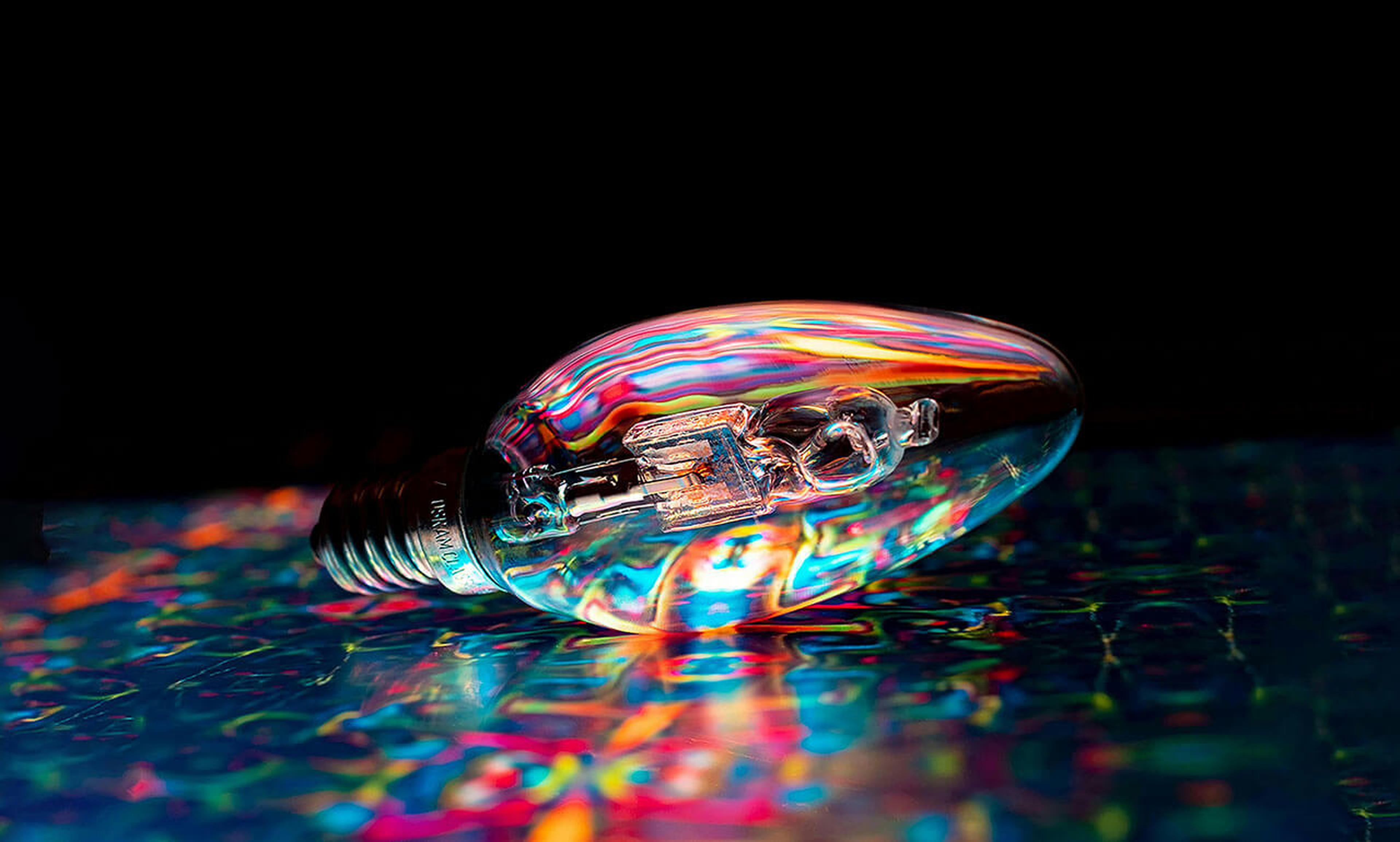 Eine Glühbirne liegt auf einer bunten, reflektierenden Oberfläche und erzeugt vor einem dunklen Hintergrund lebhafte Regenbogenmuster.