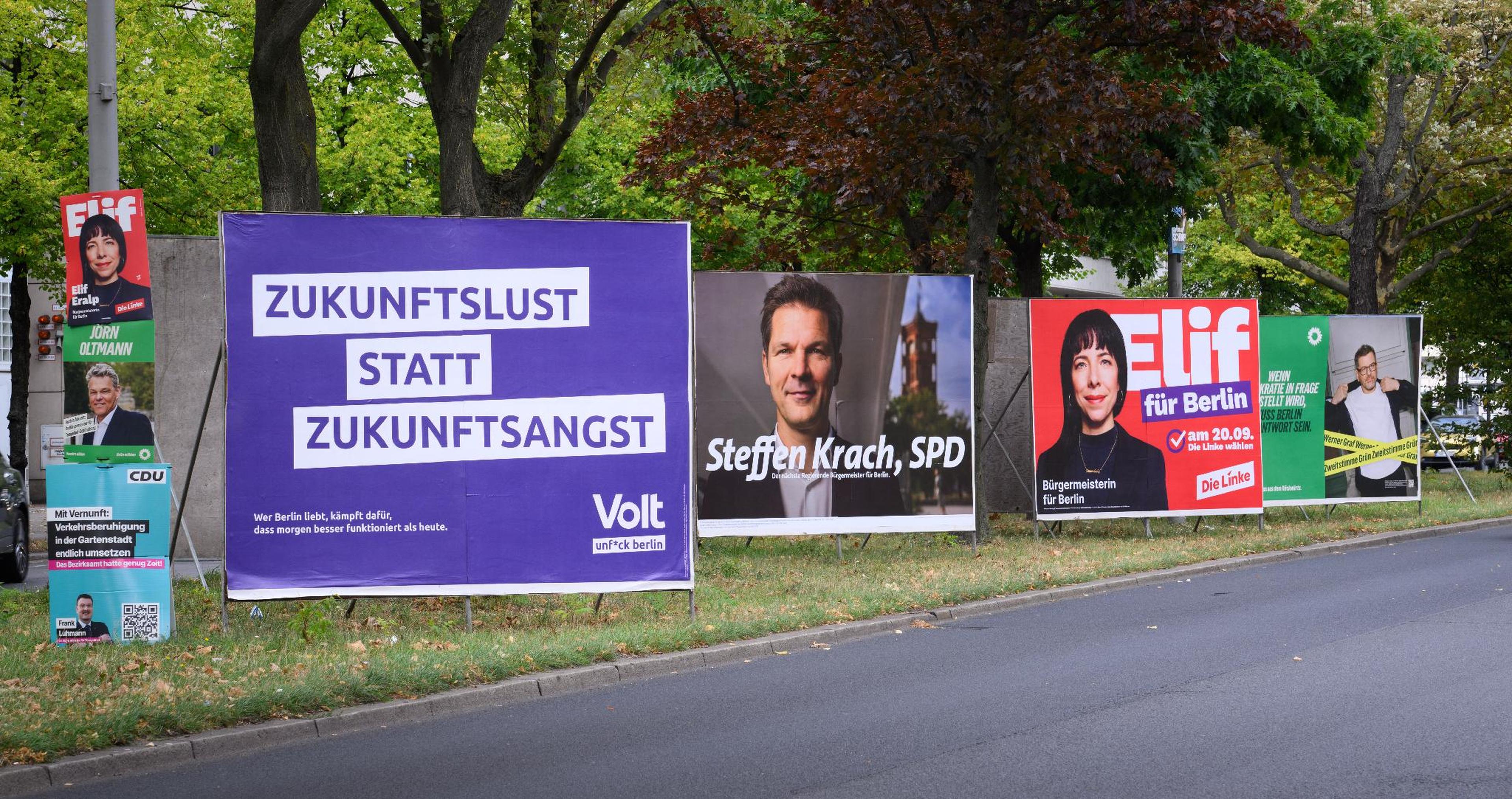 Wahlplakate säumen eine Straße; darauf sind verschiedene Kandidaten und Parolen der Parteien zu sehen, umgeben von grünen Bäumen.