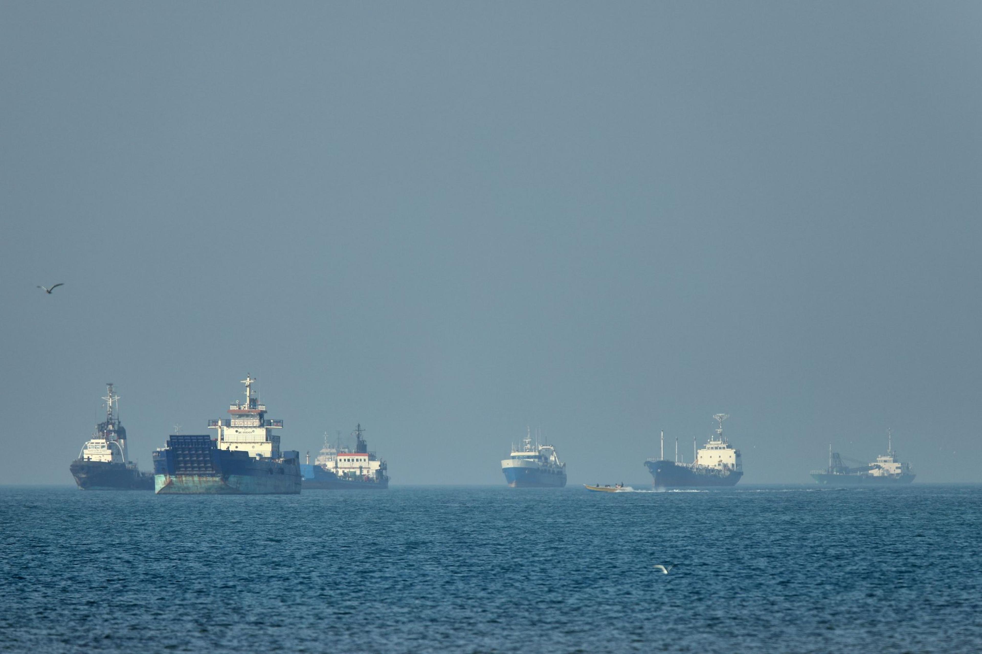Frachtschiffe, darunter Massengutfrachter und Stückgutfrachter, liegen vor der Küste vor Anker, während im Vordergrund ein kleines Motorboot in der Straße von Hormuz vor Bandar Abbas, Iran, vorbeifährt.