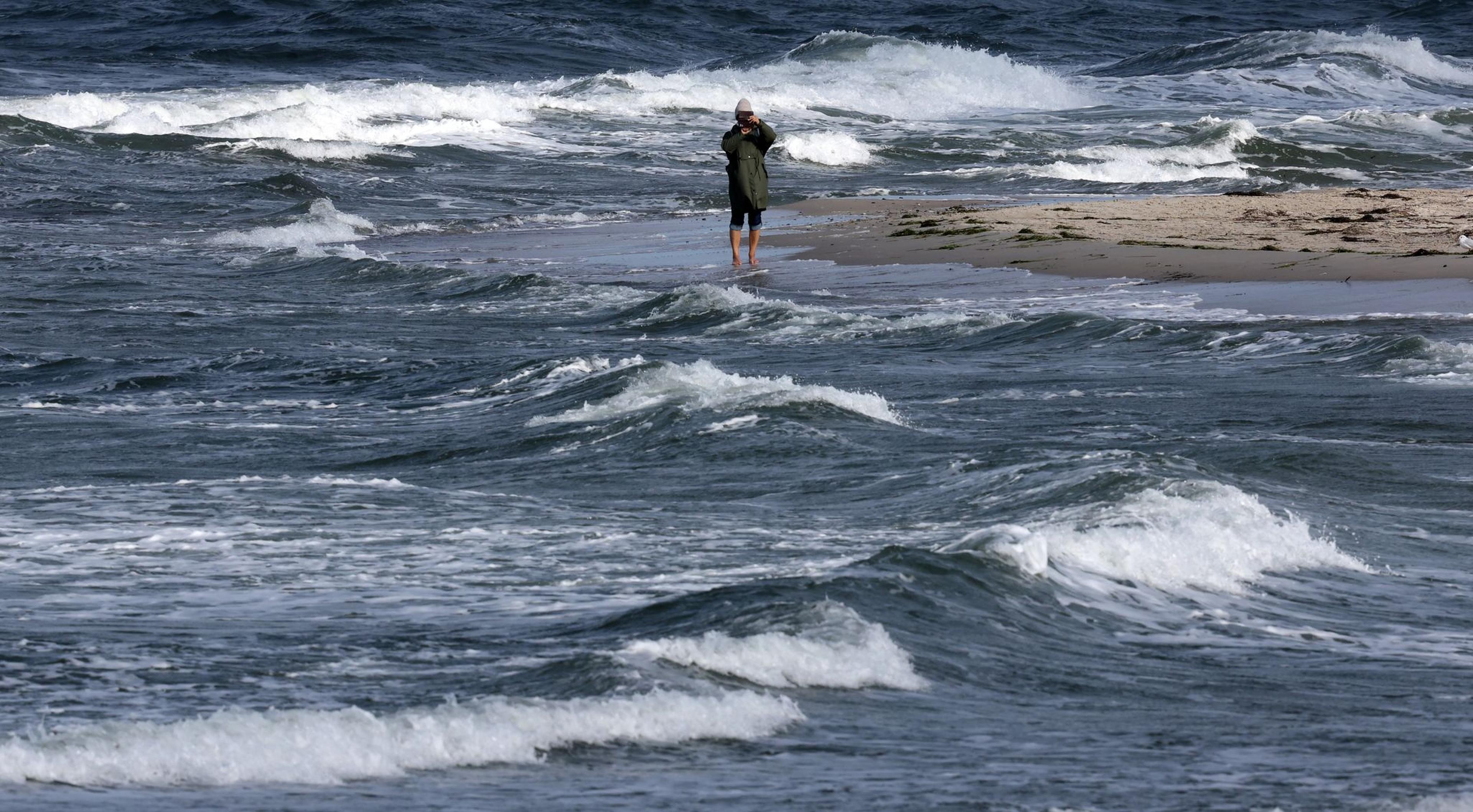 Ein Spaziergänger ist an der aufgewühlten Ostsee unterwegs. Stürmische Böen sorgen für Wellen und Gischt, die Sonne lässt das Wasser leuchten.