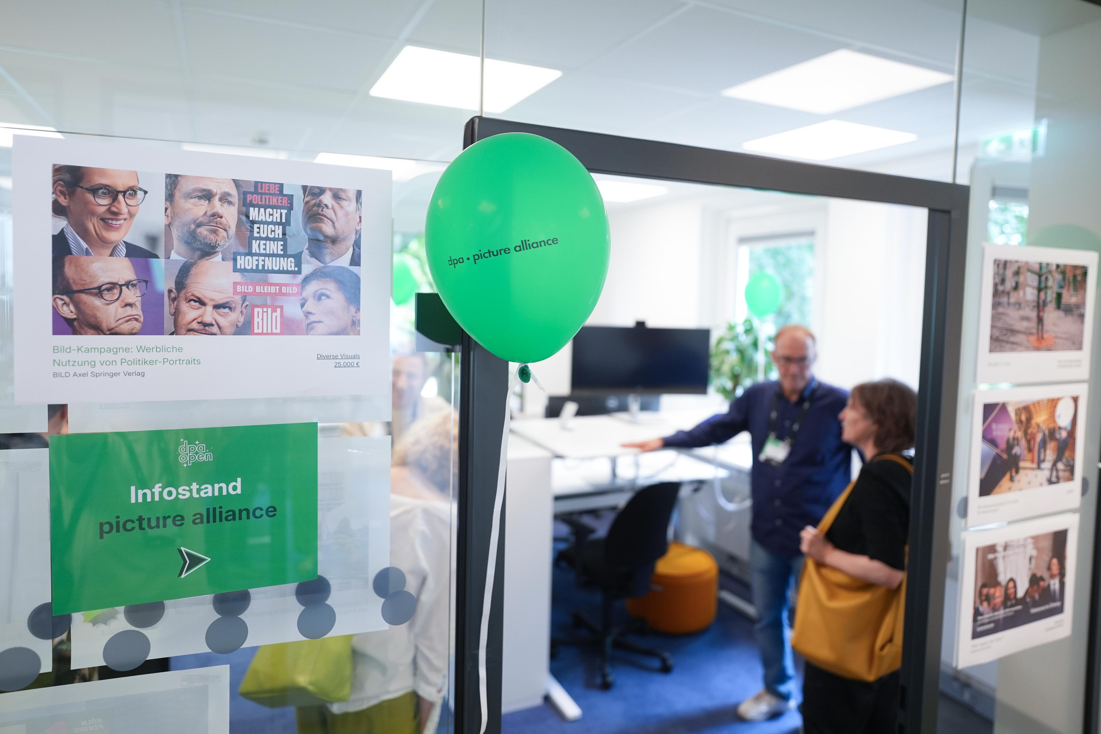 Eingang zu einem Büro mit Plakaten, grünen Luftballons und zwei Personen, die sich drinnen unterhalten. Auf einem Schild steht „Infostand picture alliance“.