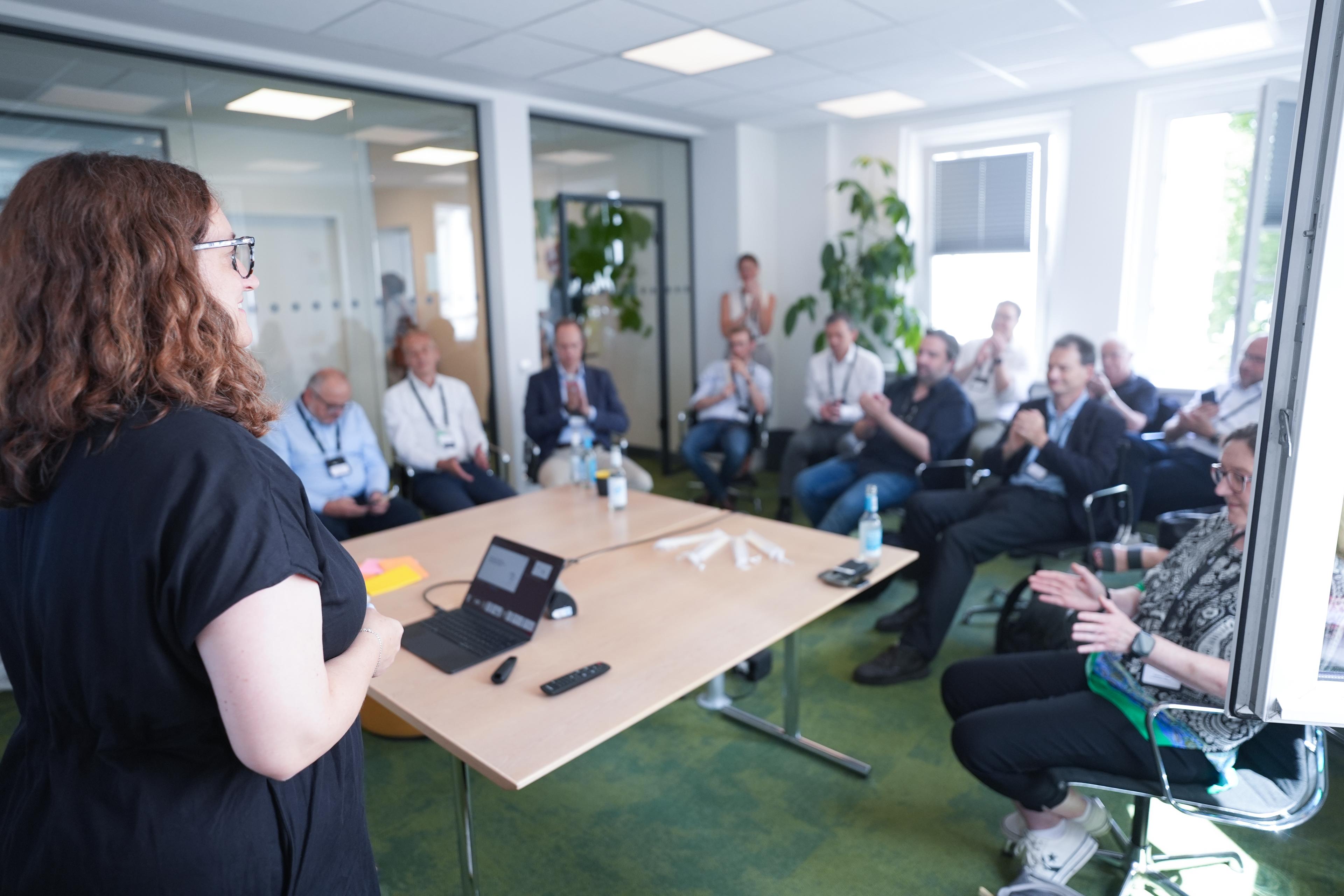 Eine Person hält in einem hell beleuchteten Konferenzraum vor einem sitzenden Publikum einen Vortrag. Auf dem Tisch liegen ein Laptop und Notizen.