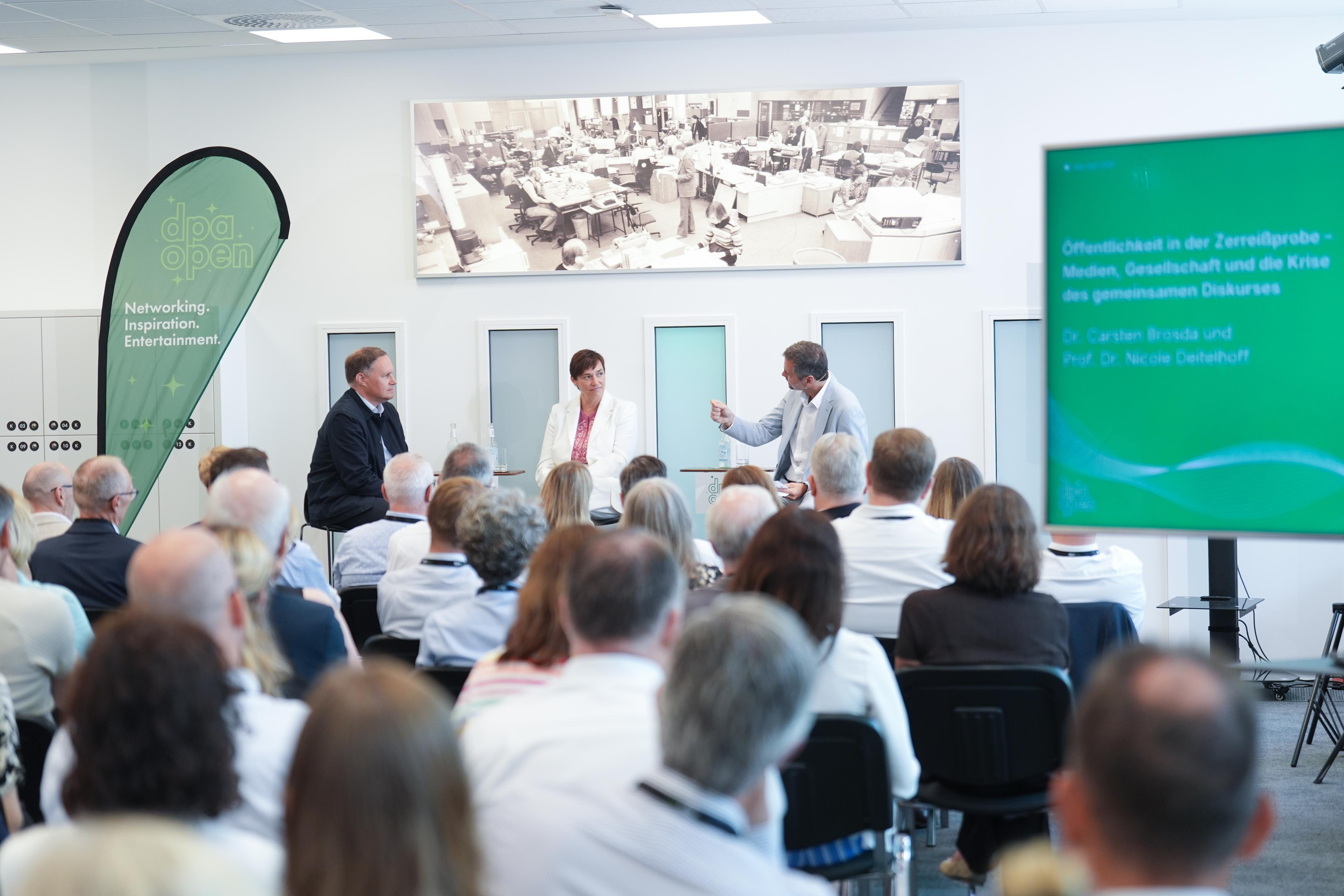 Eine Podiumsdiskussion mit drei Teilnehmern auf der Bühne und einem anwesenden Publikum, was von hinten fotografiert ist