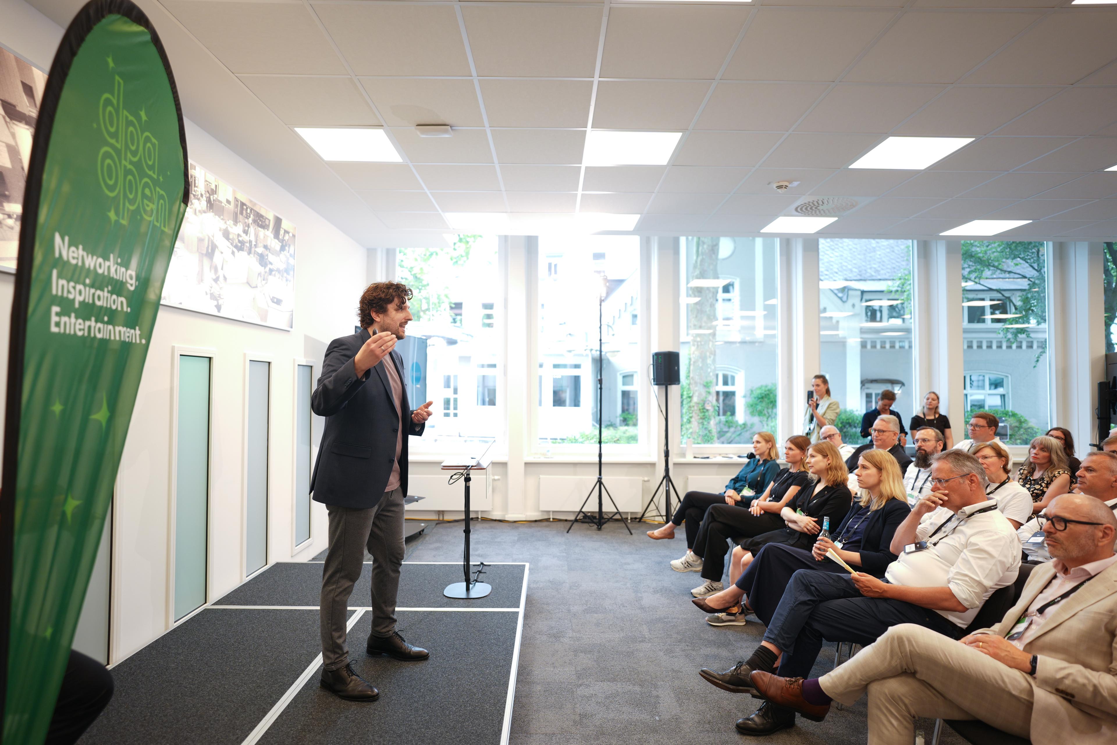 Ein Mann hält auf einer kleinen Konferenz in einem Saal eine Rede, im Hintergrund sitzt das Publikum neben einem grünen Banner. Durch große Fenster sind draußen Bäume zu sehen.