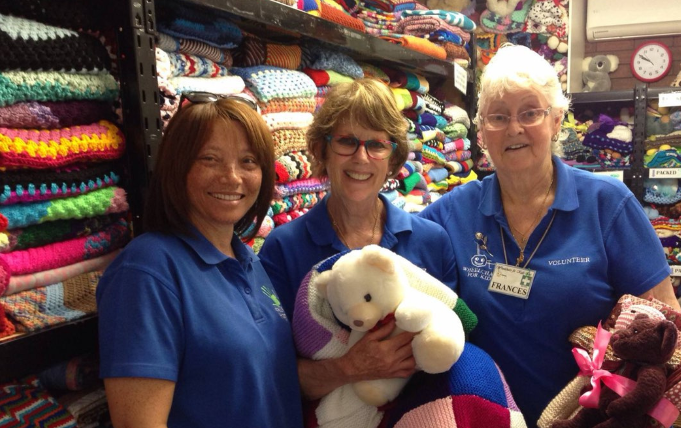 Three women in blue volunteer shirts stand smiling in front of shelves filled with colorful knitted items, holding a stuffed bear and knitted pieces.