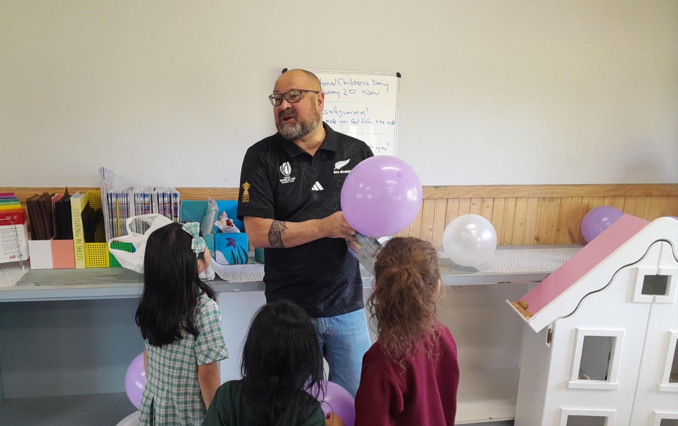A man with a beard engages with three children, holding a purple balloon in a classroom setting adorned with books and a small whiteboard.
