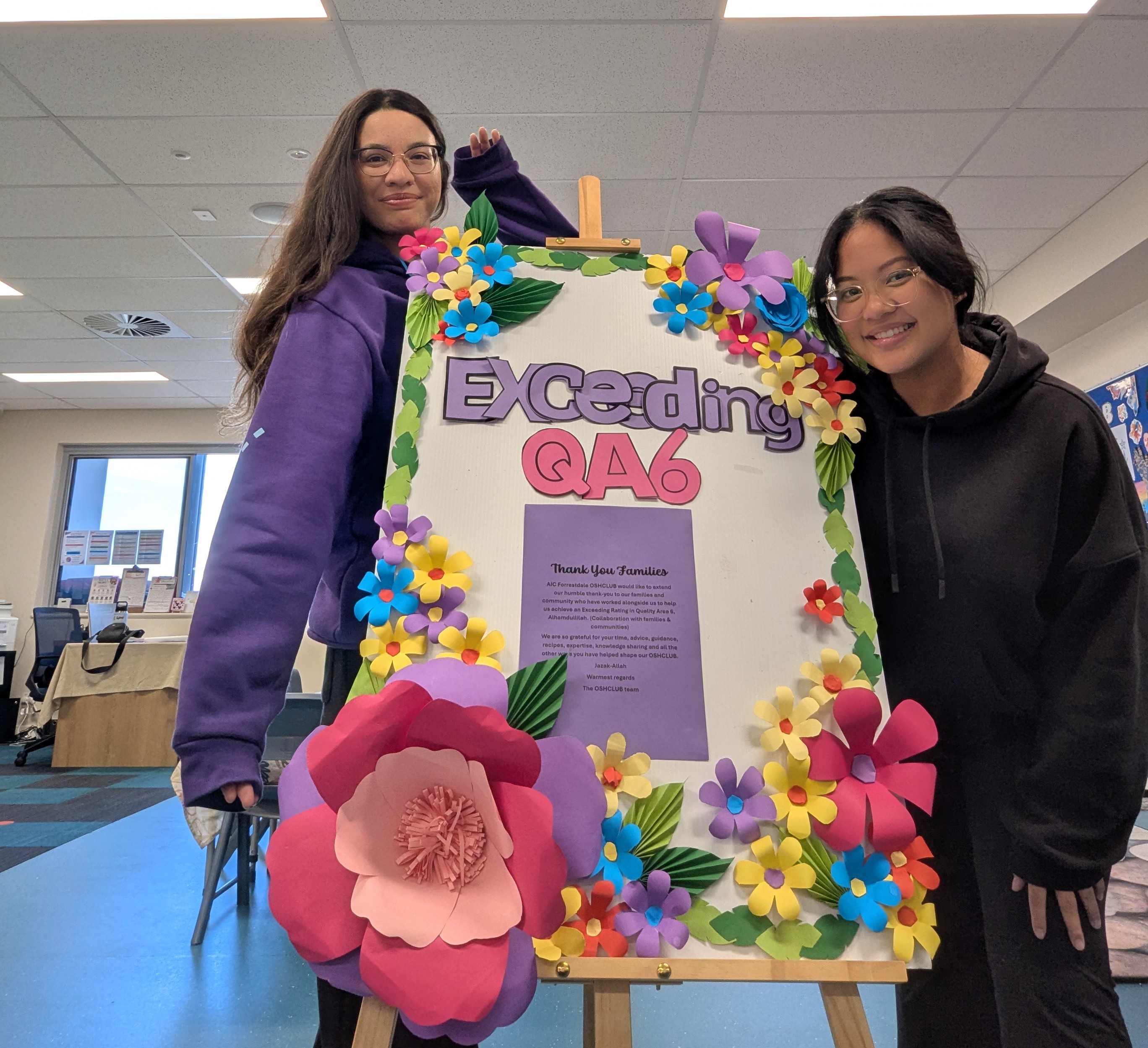 Two people smile next to a colorful floral poster on an easel that reads, "Exceeding QA6," in a room with desks in the background.