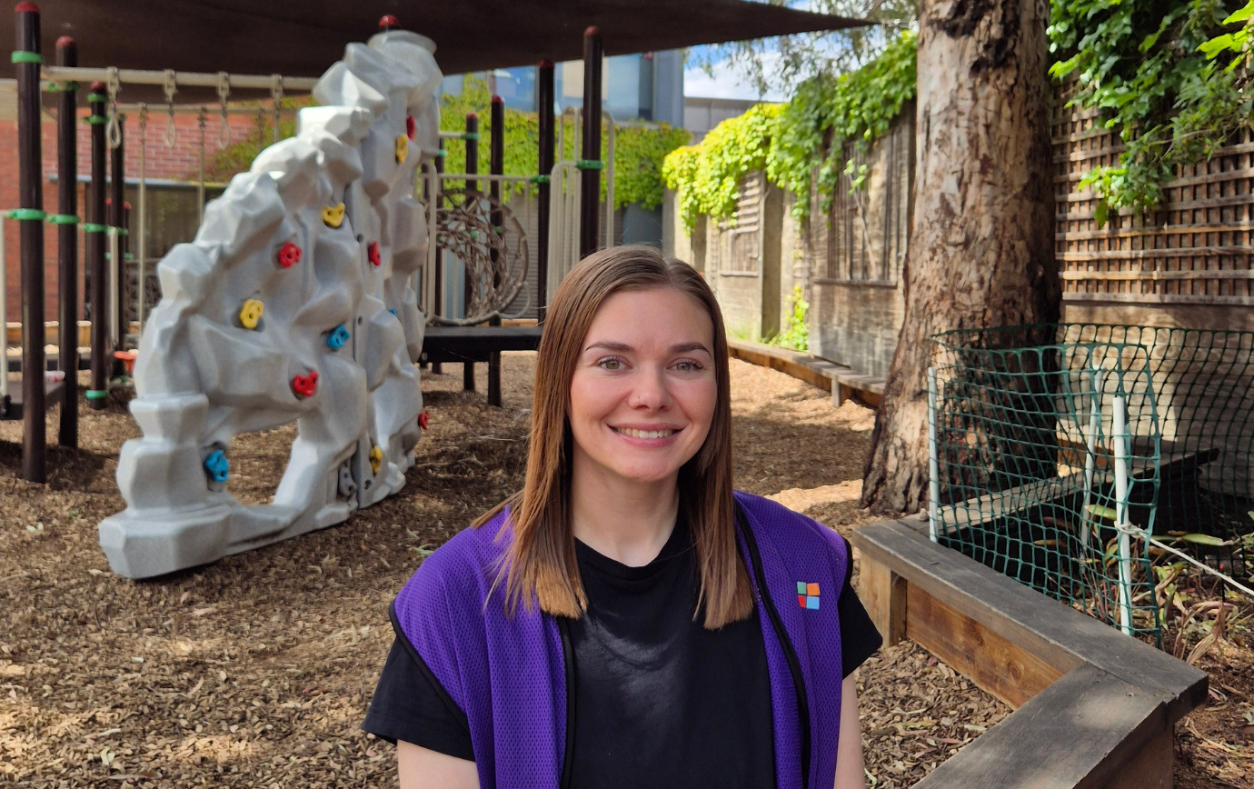 A woman in a purple vest sits smiling in a playground with a climbing structure, trees, and a netted fence in the background.