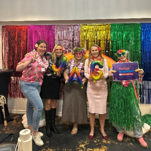 Five people in colorful outfits and accessories pose in front of a rainbow backdrop, holding festive signs and balloons.