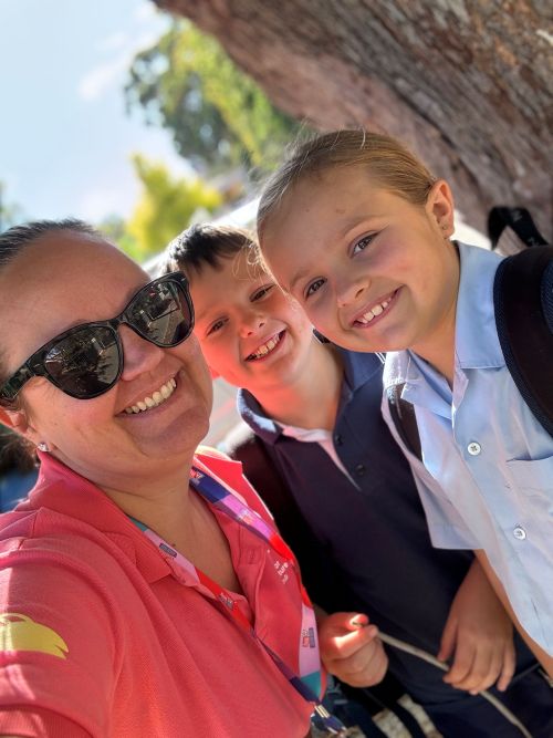 A woman wearing sunglasses takes a selfie with two smiling children in school uniforms, outdoors near a tree on a sunny day.