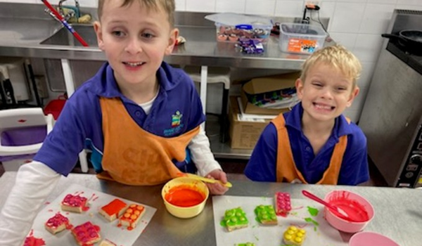 Two children in aprons decorate cookies with colorful icing at a kitchen counter, smiling and enjoying the activity.
