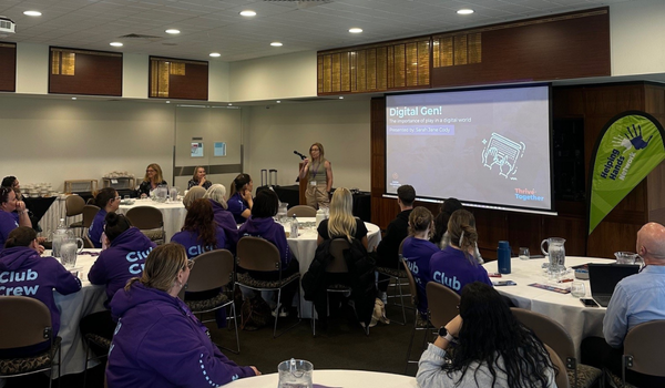 A woman presents to a group in a conference room. Attendees in purple hoodies listen and watch a presentation on a large screen.