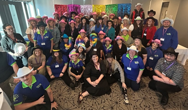 A large group of people wearing colorful hats and name tags, posing indoors in front of a vibrant, multicolored backdrop.