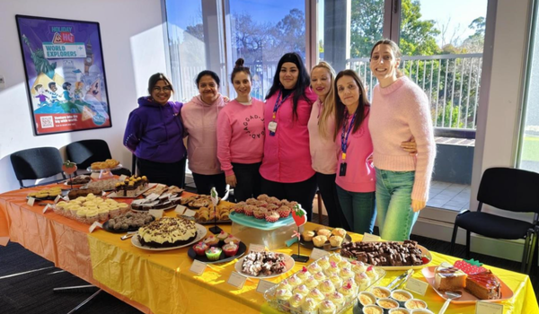 Seven people stand behind a table filled with assorted pastries and desserts, smiling for the camera in a bright room.