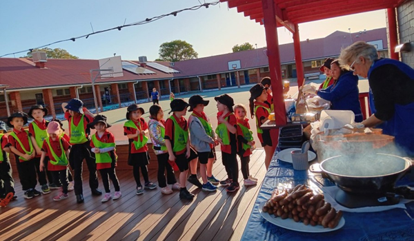 Children in bright vests line up outdoors for food served by adults at a table. A schoolyard with basketball hoops is in the background.