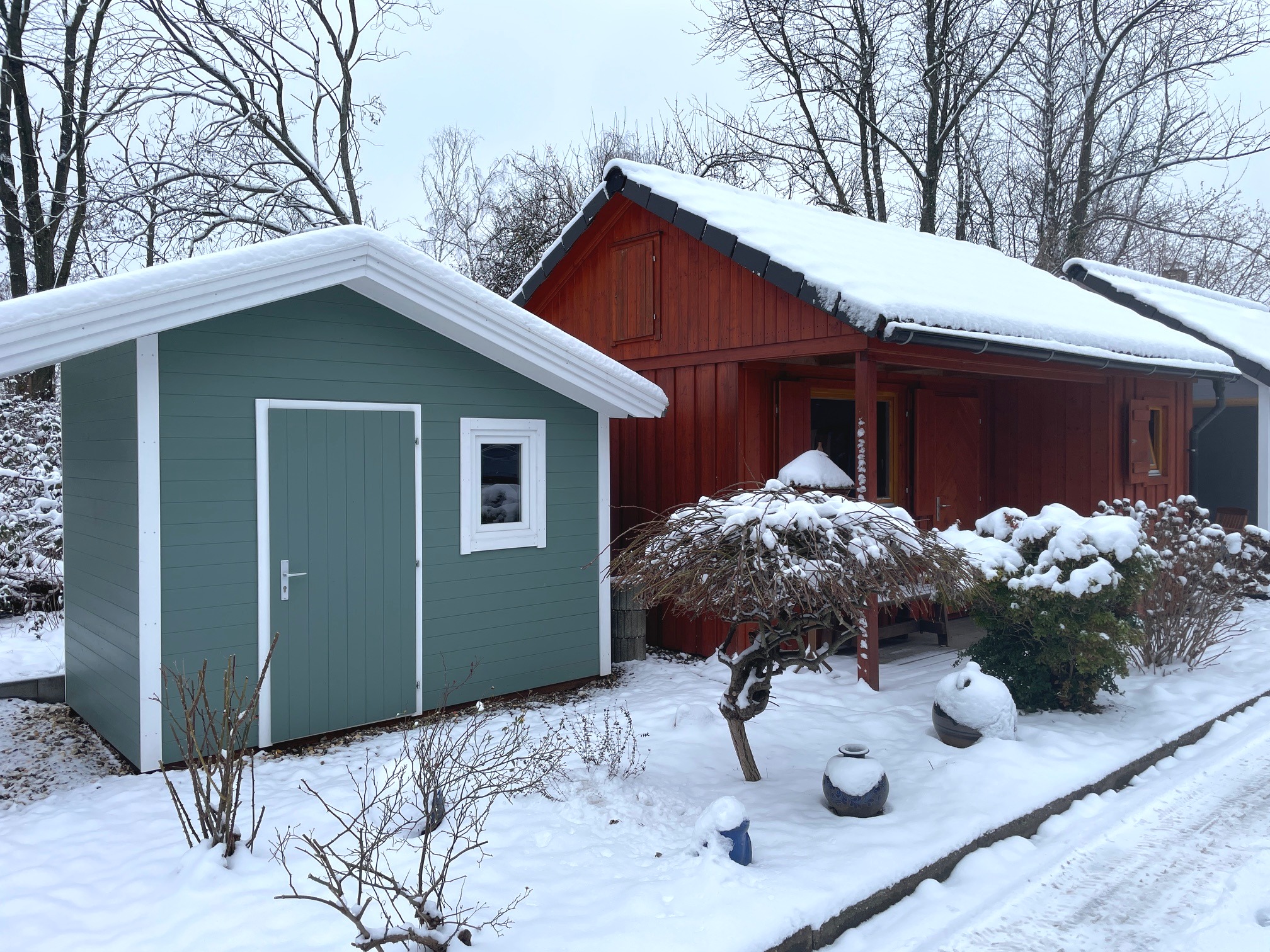 Snow-covered green shed and red wooden house surrounded by bare trees, with snow-laden bushes and a path in front.