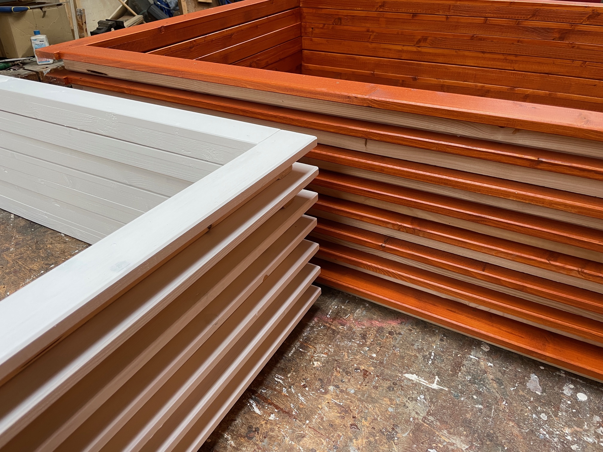 Wooden planters in progress, featuring a white and an orange-stained one, placed on a workshop floor surrounded by tools.
