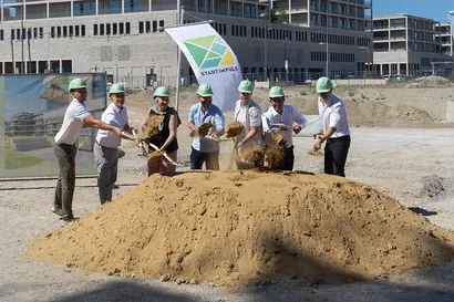 Sechs Personen mit Schutzhelmen nehmen an einem Spatenstich auf einer Baustelle teil und schaufeln Erde. Im Hintergrund sind Gebäude zu sehen.