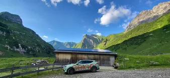 Ein Auto mit dem Schriftzug 'Geiger' steht neben einem Holzgebäude mit Solarpaneelen, in einer üppigen, bergigen Landschaft unter einem blauen Himmel mit Wolken.