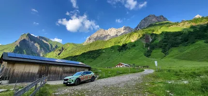 Eine malerische Berglandschaft mit einem Holzgebäude, Solarpaneelen, einem geparkten Auto, einem Kiesweg und leuchtend grünen Hügeln unter einem klaren blauen Himmel.