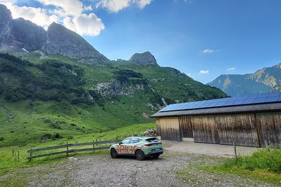 Ein Auto, das nahe einer Holzhütte mit Solarpaneelen geparkt ist, vor einer Kulisse aus üppig grünen Hügeln und felsigen Bergen unter einem klaren blauen Himmel.