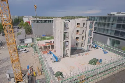 Eine Baustelle mit einem teilweise gebauten mehrstöckigen Gebäude, umgeben von Gerüsten und Kränen, in einem städtischen Gebiet unter einem bewölkten Himmel.
