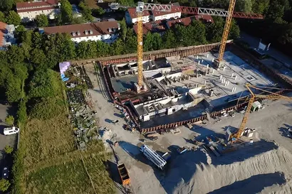 Luftaufnahme einer Baustelle mit zwei hohen Baukränen, umgeben von Wohngebäuden und Grünflächen. Die Aufnahme bietet einen umfassenden Überblick über den Baufortschritt und zeigt die Einbindung des Projekts in die umliegende Nachbarschaft.