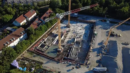 Luftaufnahme einer Baustelle mit Baukränen und Fundamentarbeiten. Das Baufeld ist von Bäumen umgeben, während sich im Hintergrund Wohngebäude befinden. Die Aufnahme vermittelt einen Überblick über den Baufortschritt und die Einbettung des Projekts in die bestehende Umgebung.