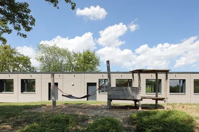 Ein modernes, niedriges Gebäude mit großen Fenstern, umgeben von Bäumen. Im Vordergrund hängt eine Hängematte zwischen zwei Holzpfosten.