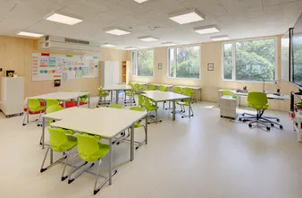 Ein hell beleuchtetes Klassenzimmer mit grünen Stühlen, weißen Tischen und großen Fenstern. An der Wand hängt eine Tafel und es gibt Poster, und ein Lehrerpult steht am Fenster.