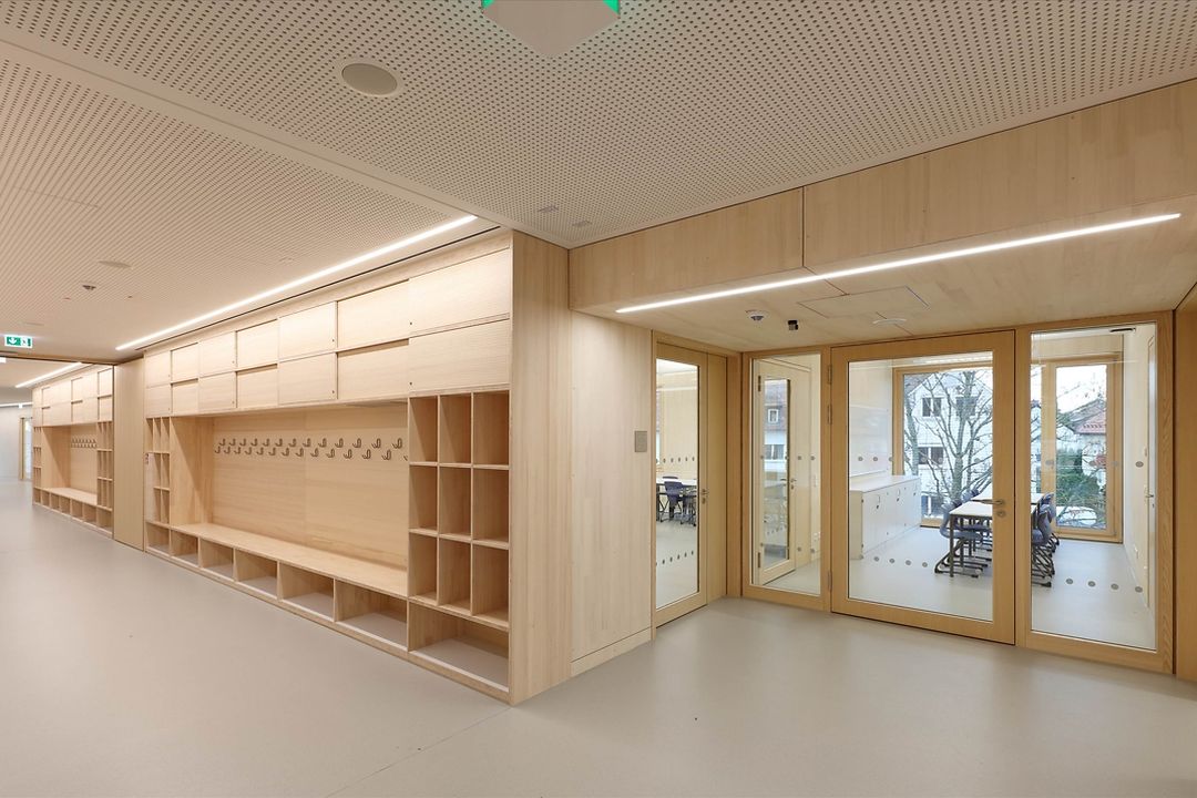 Innenansicht einer modernen Schule mit sichtbarer Garderobenanlage mit offenen Fächern, Sitzbank und Haken im Flur, daneben verglaste Klassenzimmer mit natürlicher Belichtung und hellem Holzinterieur