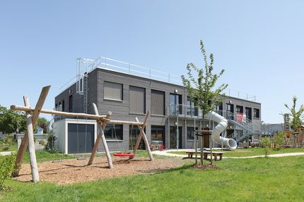 Kita-Gebäude aus Holzmdulen mit einem Spielplatz im Vordergrund.