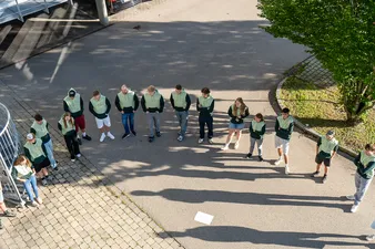 Eine Gruppe von Leuten in passenden grünen und schwarzen Hoodies steht auf einer sonnigen, gepflasterten Fläche in einem Halbkreis und wirft lange Schatten.