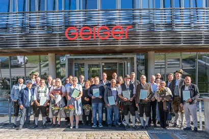 Gruppe von Leuten, die Zertifikate und Blumen halten, stehen vor einem Gebäude mit einem "Geiger"-Schild.